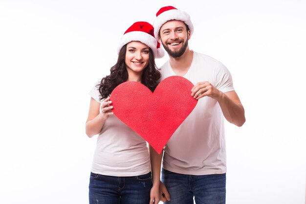 かなり若いサンタカップルの友人の男性女性のクリスマスの帽子は、白い背景のスタジオで隔離の大きな空の空白の心を保持します。明けましておめでとうございますお祝いメリーホリデーコンセプト