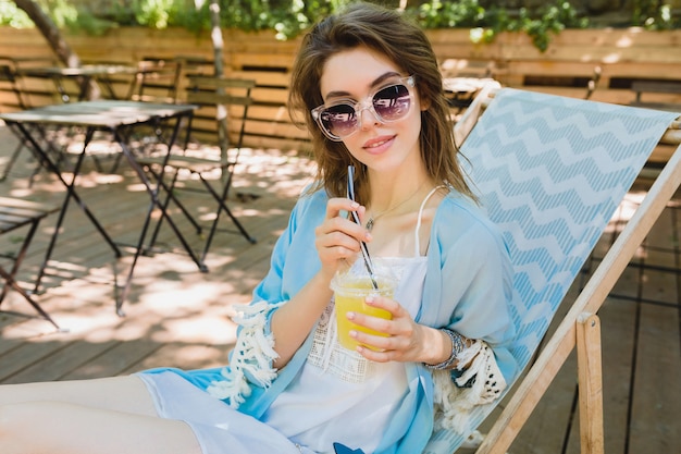 꽤 젊은 미소 세련 된 여자 여름 복장에 갑판의 자에 앉아 흰 드레스, 블루 케이프, 선글라스, 지갑을 입고, 신선한 주스를 마시고, 휴식