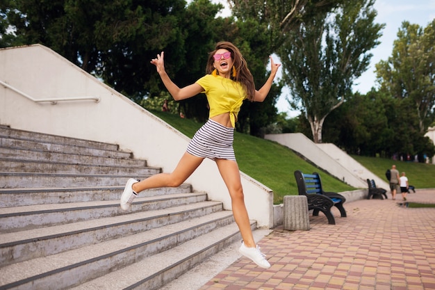 Довольно молодая улыбающаяся женщина веселится в городском парке, прыгает по лестнице, позитивная, эмоциональная, в желтом топе, полосатой мини-юбке, розовых солнцезащитных очках, белых кроссовках, тренд летней моды