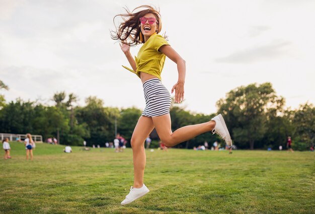 Pretty young smiling woman having fun in city park, jumping on stairs, positive, emotional, wearing yellow top, striped mini skirt, pink sunglasses, white sneakers, summer style fashion trend