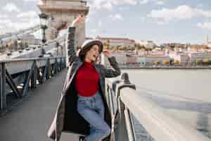 Free photo pretty young woman in blue jeans and long coat dancing on bridge near triumphal arch