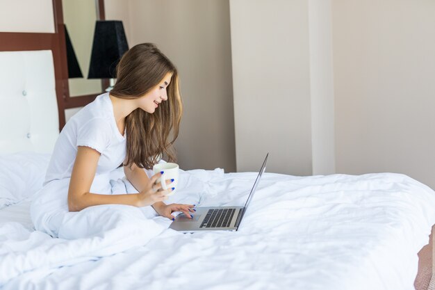 ベッドに座ってカメラに微笑んでいる間、コーヒーを飲み、ラップトップでインターネットをサーフィンしているかなり若い女性