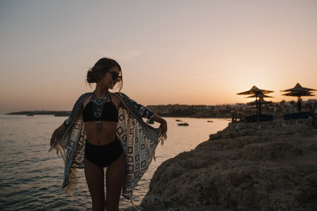 Довольно молодая женщина, наслаждаясь закатом на пляже со скалами, имея отпуск. Ловлю сторону. В стильных солнцезащитных очках, модном черном купальнике, бикини, кардигане, накидке с орнаментом.