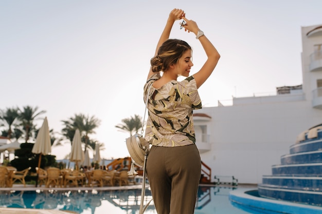 Pretty young woman enjoying time near beautiful pool, having fun in spa hotel, resort, vacation, holiday, dancing with hands up. Wearing stylish t-shirt, gray casual pants. View from back