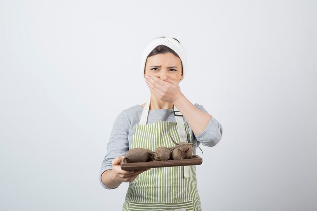 pretty young woman model in apron with a wooden board of beetroots covering mouth .