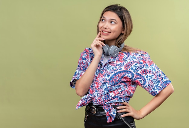 A pretty young woman in paisley printed shirt wearing headphones smiling and thinking on a green wall