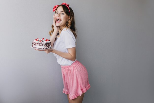 Pretty young woman in pink skirt celebrating birthday. Enthusiastic dark-haired girl dancing with sweet cake.