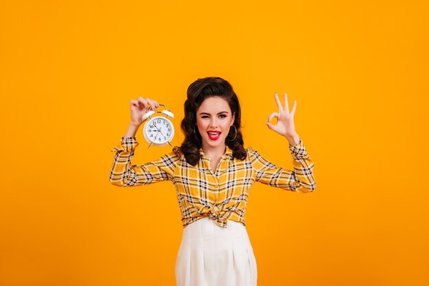 Pretty young woman posing with clock and okay sign. Smiling pinup girl in checkered shirt standing on yellow background.