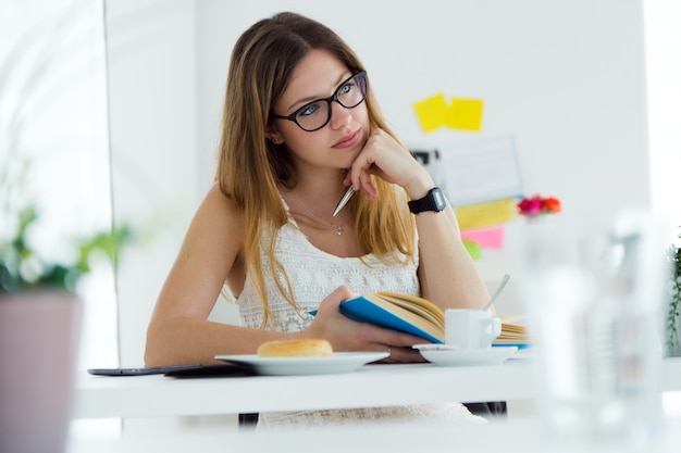 Pretty young woman reading a book and having breakfast at home.