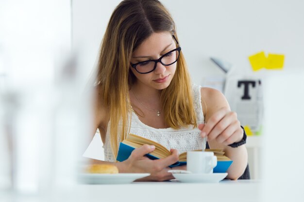 Pretty young woman reading a book and having breakfast at home.