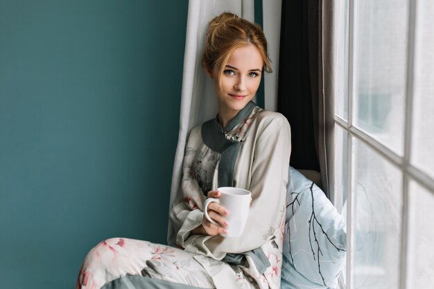 Pretty young woman sitting on window sill with mug of coffee, tea in her hand, morning relax. Wearing silk pajamas in flowers, light makeup, pure beauty.