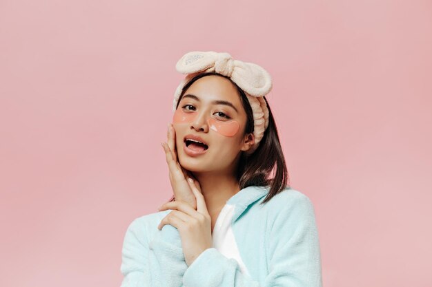 Pretty young woman in soft blue pajamas and beige headband touches her face looks into camera and poses with cosmetic eyepatches