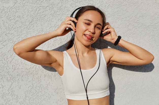Довольно молодая женщина в спортивной одежде с наушниками, слушая музыку во время тренировки на открытом воздухе, глядя в камеру, показывая язык и подмигивая, имеет забавное выражение лица.