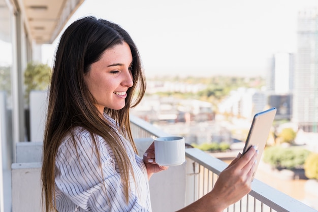 Pretty young woman standing in balcony making video call on smartphone holding coffee cup in hand