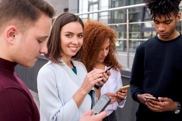 Pretty young woman standing between her friends using mobile phones looking at camera