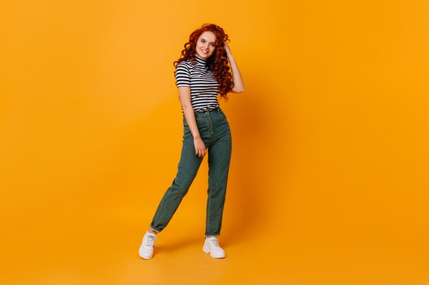 Pretty young woman touches her curly red hair and smiles. Portrait of girl in jeans and top in orange studio.