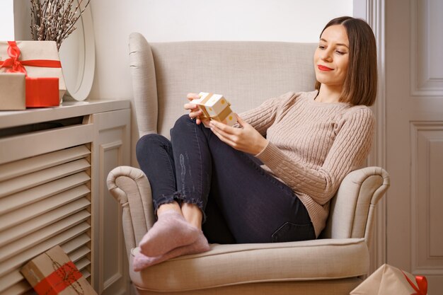 PRETTY young woman while sitting in a comfortable armchair holding a gift box surrounded by gifts at home.