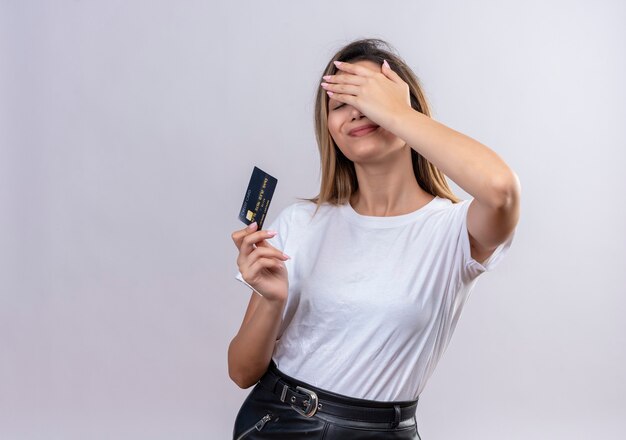 A pretty young woman in white t-shirt showing credit card while keeping hand on forehead on a white wall