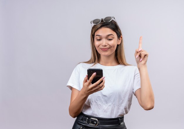 A pretty young woman in white t-shirt wearing sunglasses on her head pointing with index finger while looking at mobile phone on a white wall