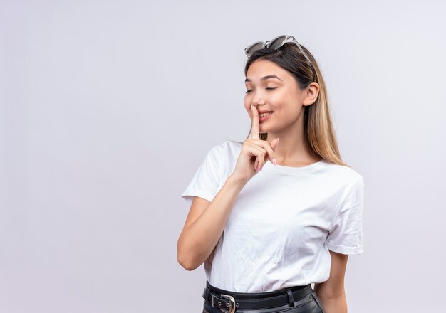 頭にサングラスをかけた白いTシャツを着たかなり若い女性が白い壁に目を閉じてshhジェスチャーを示しています