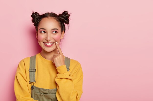 Pretty young woman with Asian appearance, wears yellow sweatshirt and overalls, has two hair buns, looks aside