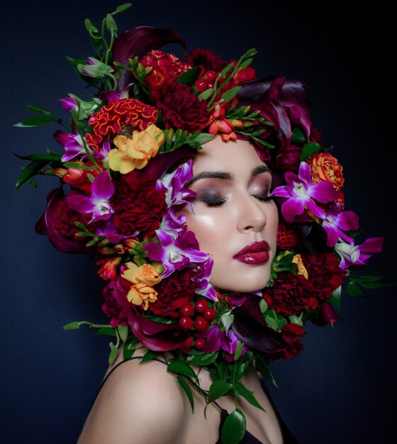 Pretty young woman with bright make-up with closed eyes surrounded with colorful wreath made of fresh flowers on the dark blue background