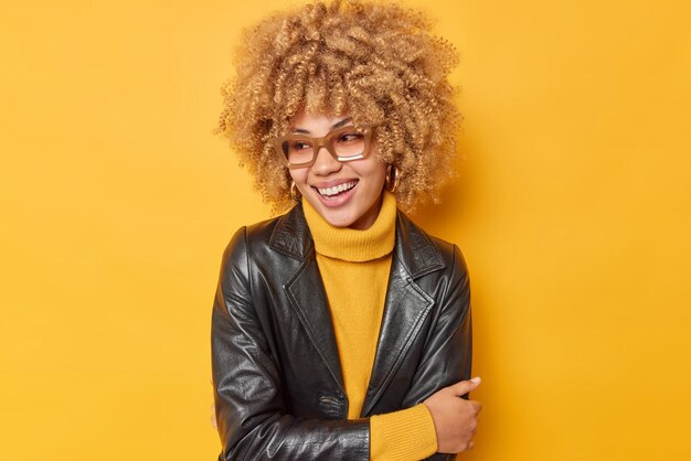 Pretty young woman with curly bushy hair looks away happily being in good mood wears spectacles warm jumper and leather jacket isolated over yellow background. People and happy emotions concept