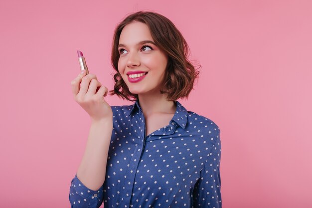 Pretty young woman with curly hair isolated on pink wall with lipstick in hand. smiling lovable girl wears blue blouse.