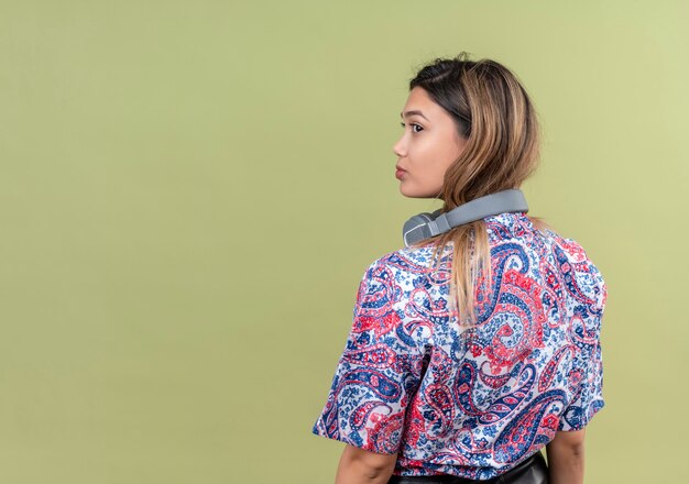 A pretty young woman with long hair in paisley printed shirt wearing headphones looking far away on a green wall