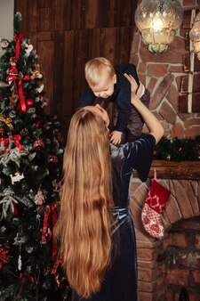 Довольно молодая женщина с годовалым ребенком играет у елки в гостиной. мама с милым сыном в комнате, украшенной рождеством. они улыбаются и счастливы. концепция семейного нового года. копировать пространство