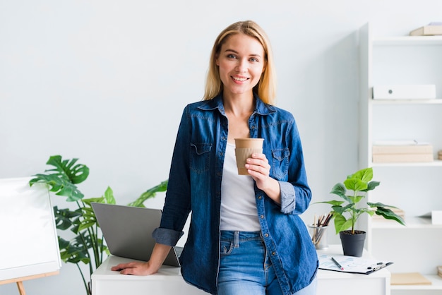 Foto gratuita giovane donna graziosa con la tazza di carta nel posto di lavoro