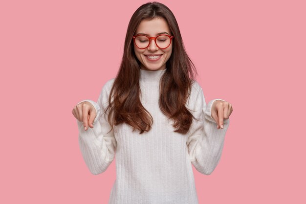 Pretty young woman with tender smile points down with both index fingers, demonstrates new shoes she bought