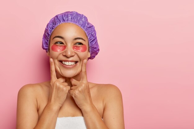 Pretty young woman wrapped in soft towel, cares about skin, wears hydrogel recovery patches or collagen pads, touches cheeks with index fingers, wears bath cap, shows bare shoulders, healthy skin