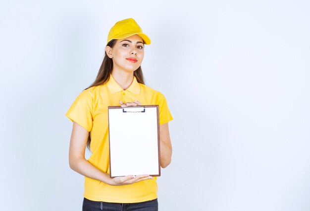 Pretty young woman in yellow courier uniform with clipboard posing over white wall.