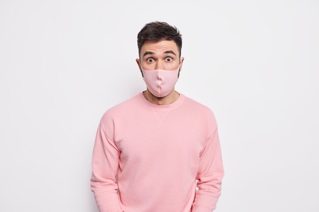 Free photo prevention and safety concept. surprised young man wears protective mask on face prevents coronavirus spread finds out shocking statistics dressed in pink sweater