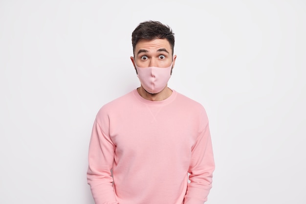 Prevention and safety concept. Surprised young man wears protective mask on face prevents coronavirus spread finds out shocking statistics dressed in pink sweater 