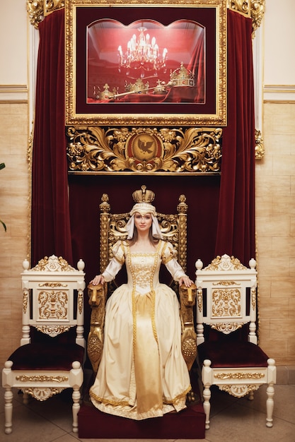 Princess in rich golden dress sits on the throne before red wall