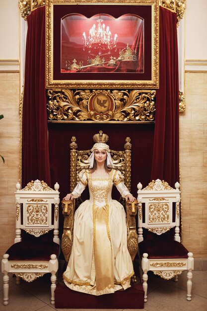 Принцесса в богатом золотом платье сидит на троне перед красной стеной