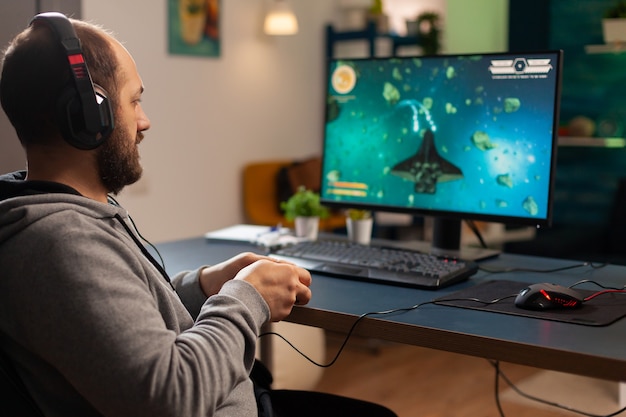 Pro man gamer playing video game at professional computer late night wearing headphones. Excited player using wireless controller for virtual tournament gaming space shooter at home