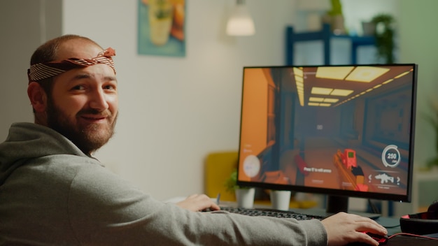サイバースペースでのゲーム選手権中にFPSシューティングビデオゲームをプレイしてカメラの笑顔を見て頭を回すプロの男のゲーマー。オンライントーナメントで強力なコンピューターで実行するeスポーツプレーヤー