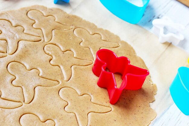 Process of dealing with gingerbread man cookies, use red gingerbread man mold cutting gingerbread dough on baking paper around colorful cookie cutters on white wooden table. top view