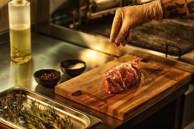 Process of preparing beef steak in professional restaurant kitchen chef in white glove salting meat using such ingredients as seasonings pepper oil and rosemary for spicy taste