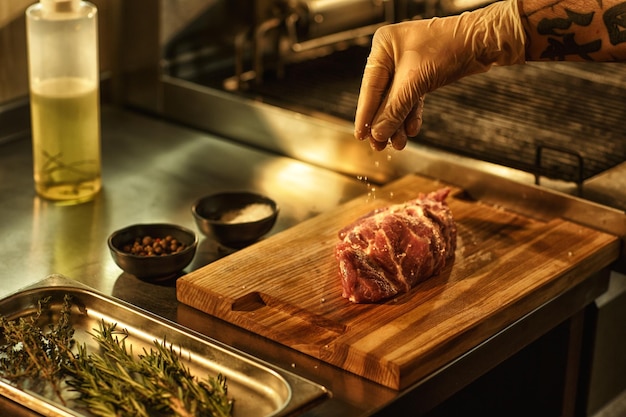 スパイシーな味わいの調味料ペッパーオイルやローズマリーなどの材料を使用して、白い手袋で肉を塩漬けにするプロのレストランのキッチンシェフでビーフステーキを準備するプロセス