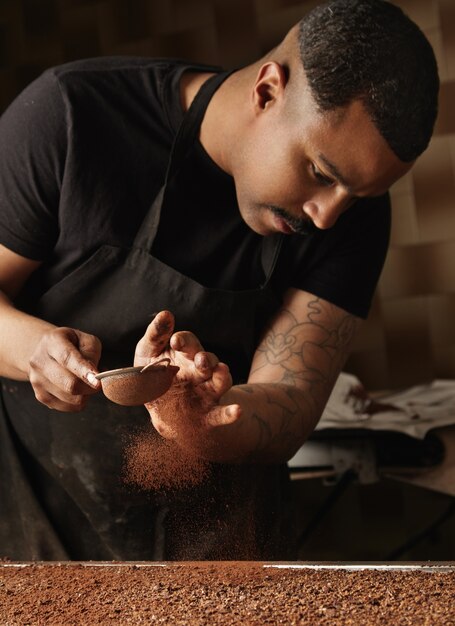 Professional baker pours cinammon on mold filled with melted chocolate mass. Preparation of tasty cake from organic chocolate in artisan confectionery for sale