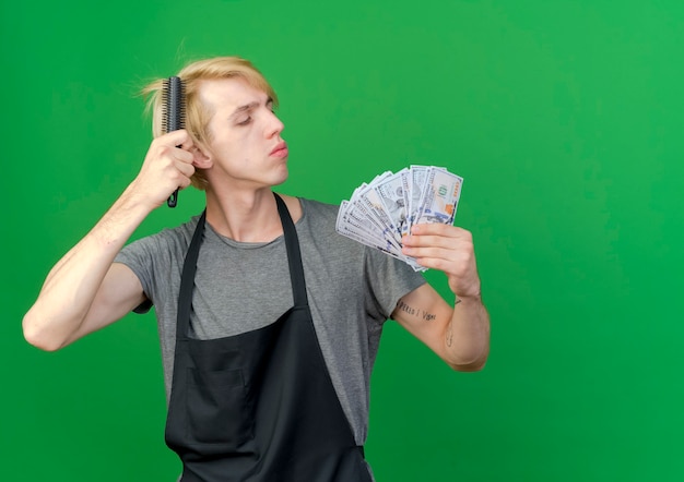 Uomo di barbiere professionista in grembiule che tiene contanti e pettine per capelli che pettina i capelli con la faccia seria