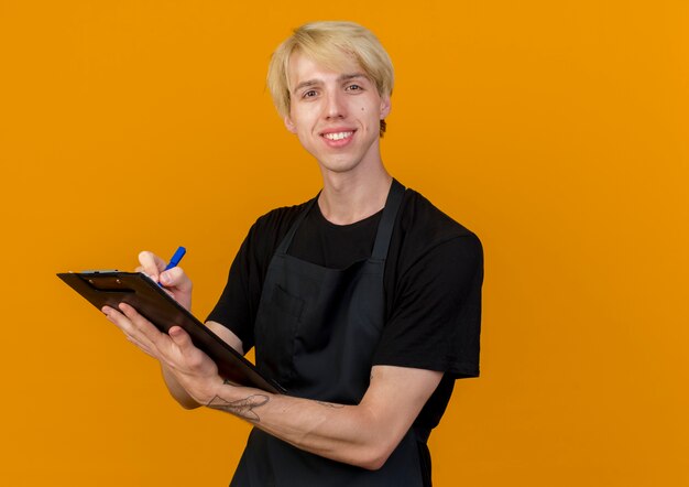 Professional barber man in apron holding clipboard and pen looking at front with smile on face standing over orange wall