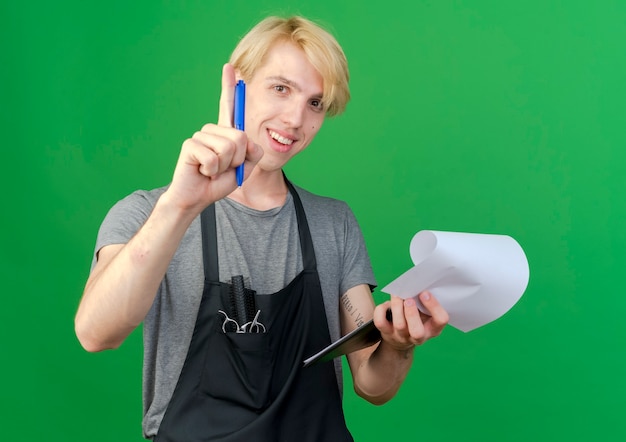 Professional barber man in apron holding clipboard with blank pages and pen smiling 