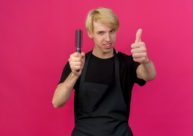 Professional barber man in apron holding hair brush smiling showing thumbs up 