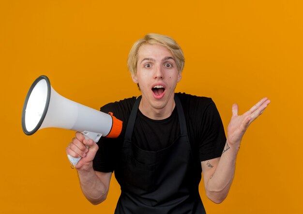 Professional barber man in apron holding megaphone looking at front being surprised and amazed standing over orange wall