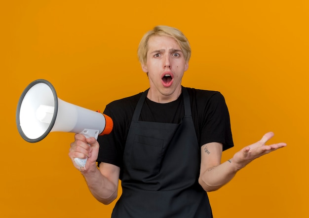 Foto gratuita uomo di barbiere professionista in grembiule che tiene il megafono guardando davanti essendo sorpreso e confuso in piedi sopra la parete arancione